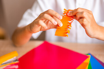 Boy's hands cut orange origami with orange scissors on multicolored paper background close-up