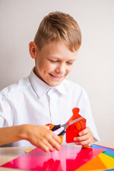 A smiling blond boy in a white cotton shirt is cutting a little man out of red paper with orange scissors