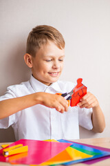 A smiling blond boy in a white cotton shirt is cutting a little man out of red paper with orange scissors