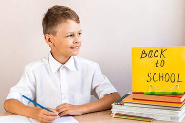 A blonde schoolboy in a white shirt sits at a desk against the background of textbooks and pencils with a yellow sign back to school