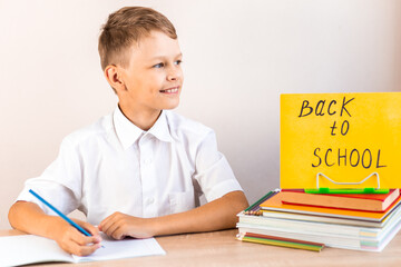 A blonde schoolboy in a white shirt sits at a desk against the background of textbooks and pencils...