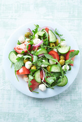 Summer Salad with Mozzarella Cheese, Cucumber, green Olives, Tomatoes and fresh Rocket. Bright wooden background. Top view. Copy space. 
