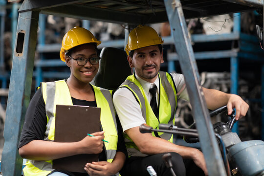African American female employee worker and hispanic man manager wearing safety hardhat helmet working together on forklift truck at old auto and car parts warehouse store.