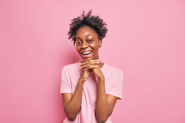 Portrait of pretty dark skinned Afro American woman keeps hands under chin smiles broadly dressed in casual t shirt isolated over pink background. People sincere emotions and happiness concept.