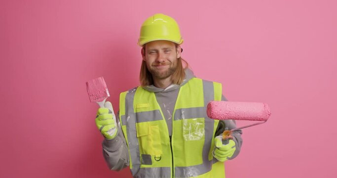 Positive carefree redhead man dressed in working clothes has fun dances with painting tools happy to finish house redecoration isolated over pink background. Repair and refurbishment concept