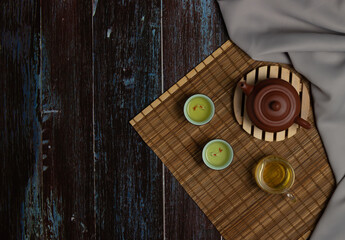 Fototapeta premium Chinese Tea Ceremony. Brown ceramic teapot and turquoise teacup. Green tea on a bamboo mat on an aged wooden table. Top view. Copy space