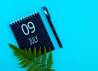 July 9th. Day 9 of month, Calendar date. Black notepad sheet, pen, fern twig, on a blue background....