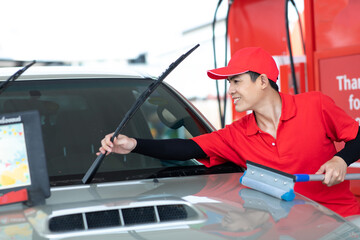 Man worker in red uniform using sponge  clean windscreen and checking wiper blade of car while...
