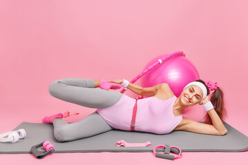 Horizontal shot of fitness woman with pony tail lies on mat stretches legs enjoys doing gymnastics wears sportsclothes has happy expression surrounded by sport equipment isolated over pink background
