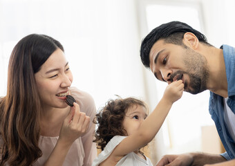 Happy moment family eating chocolate cookie in living room at home. Father Mother and daughter laughing having a good meal in the morning.