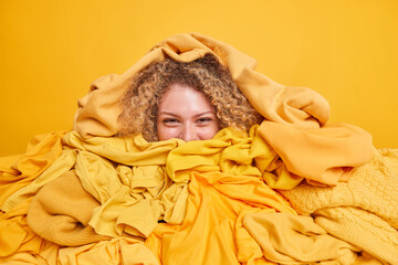 Positive curly haired woman covered with pile of clothes stays at home during quarantine sorts out...