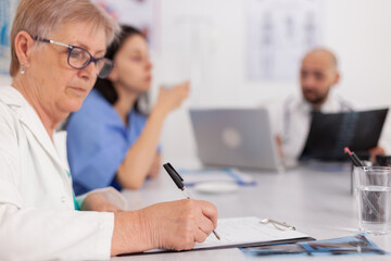 Obraz premium Physician senior woman in white coat analyzing medical examination prescribing pill medication writing sickness treatment on clipboard. Hospital teamwork sitting at desk in conference meeting room