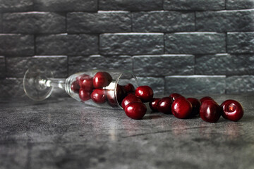 The fallen glass with scattered cherries on the table against the background of a gray brick wall