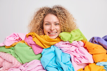 Positive good looking woman with curly hair sorts clothes from wardrobe at home demonstrates her...