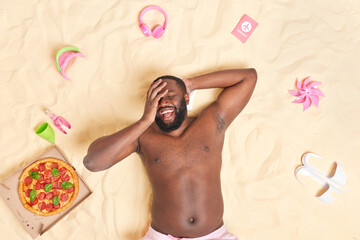 Fototapeta na wymiar Outdoor shot of happy bearded dark skinned man lies with bare torso on white sand keeps hand on face laughs joyfully surrounded by tasty pizza slippers passport headphones beach toys has upbeat mood
