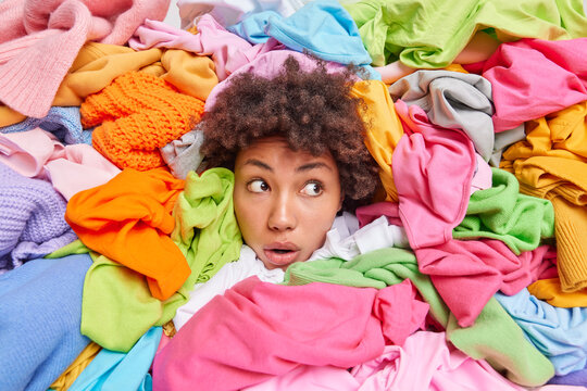 Woman head sticking out through different multicolored clothes looks away with stunned expression arranges closet purges unnecessary clothing from wardrobe. Female shopaholic poses around attire
