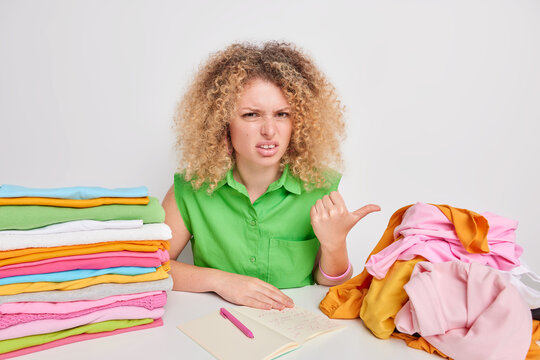 Displeased curly haired woman points with thub at unfolded laundry feels tired of domestic work makes notes in notepad writes down washing temperature for clothes made of different material.