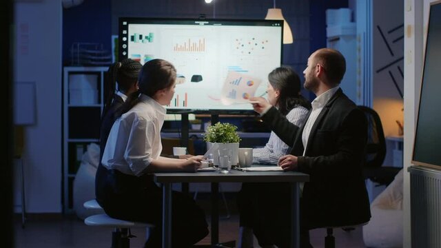 Diverse business teamwork of entrepreneur analyzing management strategy working in office room late at night. Multi ethnic businesspeople looking at company presentation on monitor sitting at table