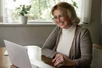 Happy senior Caucasian woman look at laptop screen talk speak on video call on device from home....