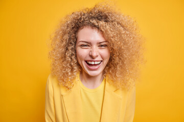Portrait of good looking curly haired young woman smiles broadly expresses positive emotions glad...
