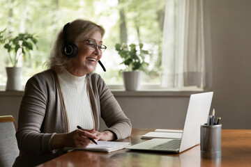 Smiling modern Caucasian old woman wear headset have fun use computer study online on gadget. Happy smart middle-aged female in earphones write note take distant course or training on laptop.