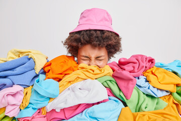 Disorganised curly haired woman sticks head out of clothes heap wears pink panama does household...