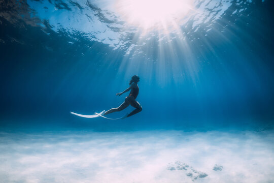 Freediver with white fins glides and posing underwater in sea with sunlight.