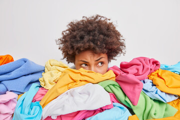 Serious Afro American woman poses near heap of multicolored unsorted clothes after washing brings...