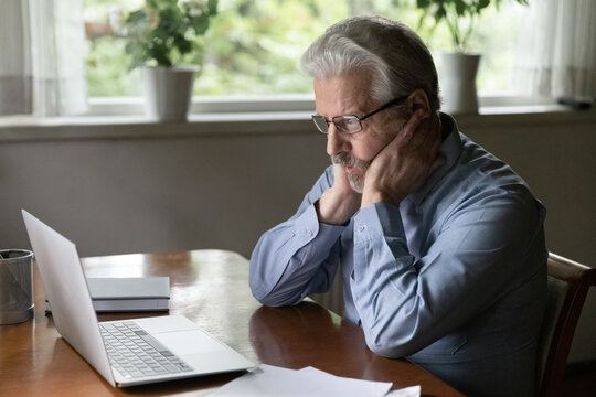 Pensive upset mature 60s Caucasian man sit at table at home work on computer with paperwork think ponder. Thoughtful elderly male use laptop look at screen make decision solve problem on device.