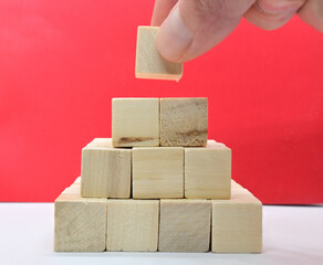 Stacked small wooden cubes on a white surface create a pyramid with a hand adding the last piece against a red background. Success concept.