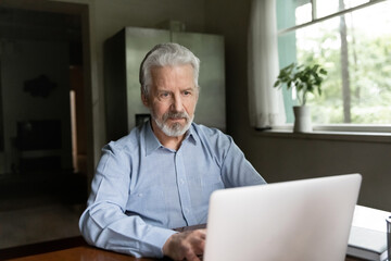 Serious mature Caucasian man sit at desk at home office look at laptop screen work online on gadget. Pensive modern smart old 70s male use computer browse internet on web. Elderly technology concept.
