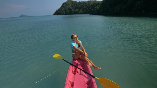 Pretty sportive blonde lady in stylish swimsuit sails on pink plastic canoe along azure ocean bay at exotic resort. Traveling to tropical countries. Girl is sailing on kayak in ocean, view from above.