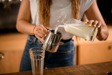 woman's hand holds steel jug and pours milk into it from bottle.