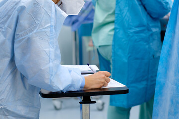 A female doctor in a blue uniform and a medical mask makes notes in documents during surgery.