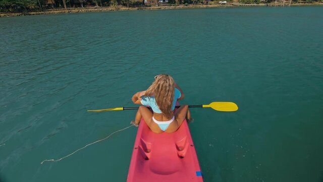 Seductive sexy blonde woman in blue swimsuit sails on pink plastic kayak along azure sea at exotic resort backside upper above view. Traveling to tropical countries. Girl is sailing on kayak in ocean.