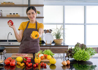 Young housewife stand smiling,  hold red and yellow bell pepper with both hands. Offering the yellow one on in the front. The kitchen counter full of various kinds of vegetables.