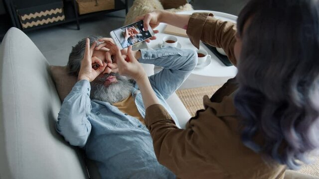 Joyful mature husband is posing for smartphone camera making funny faces and gestures while wife is taking pictures touching screen enjoying photography