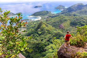 Young female traveller standing at the edge of the cliff at Morne Blanc View Point, overlooking Mahe Island coastline with lush tropical vegetation and crystal blue ocean, Seychelles. - obrazy, fototapety, plakaty