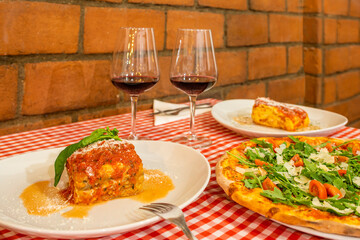 Table in the dining room of an Italian restaurant with pizza to share and individual plates of lasagna and two glasses of red wine to soak your throat