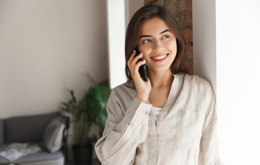 Young woman standing near office window, having phone call. Girl freelancer talking on smartphone, having conversation and smiling, answer customer, working remote