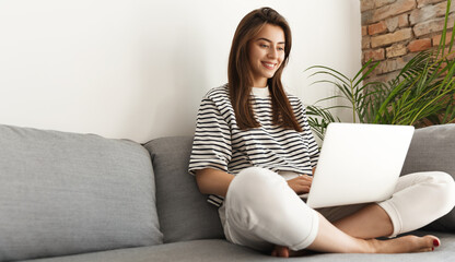 Young brunette girl student using computer from home. E-learning or remote freelance work concept. Woman working in living room with laptop, watching videos on device