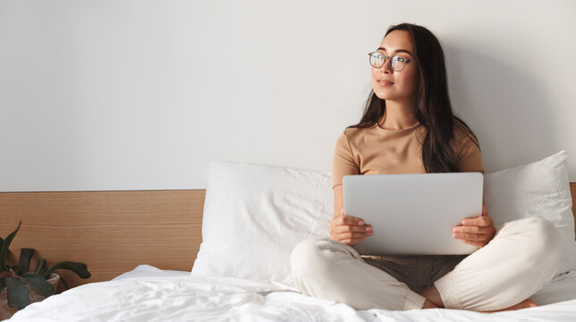Young Asian Girl Student Studying With Laptop In Her Bed. Young Woman Working Remote From Home Bedroom, Sitting In Glasses With Computer And Looking Outside, Writing Blog