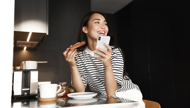 Image of happy asian woman eating breakfast and using smartphone. Girl laughing, eat toast and drink coffee, holding mobile phone in kitchen at home