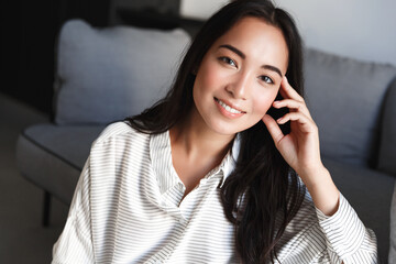 Portrait of young asian woman sitting at home in living room, Girl with glowing natural skin, no make up, smiling happy at camera, resting and relaxing on weekend inside house, leisure time