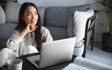 Image of asian girl studying on computer from home. E-learning during pandemic, young woman freelancer working with laptop, looking outside window and smiling, drinking coffee