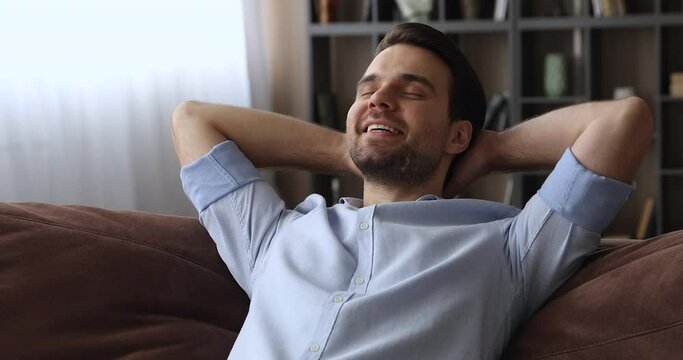Close up serene calm millennial smiling man put hands behind head lean on comfy sofa resting alone at home, feel satisfaction enjoy fresh conditioned air inside of cozy living room, relaxation concept