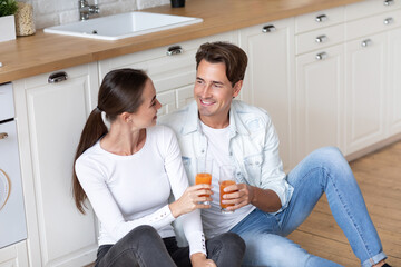 Smiling couple young guy and girl sit on warm kitchen floor talking enjoying drinking orange juice at modern home. Good weekend.