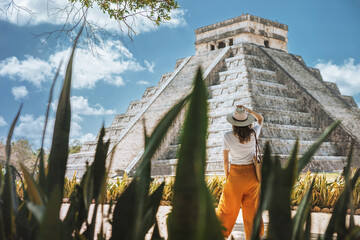 A young woman tourist in a hat stands against the background of the pyramid of Kukulcan in the ancient Mexican city of Chichen Itza. Travel concept.Mayan pyramids in Yucatan, Mexico - obrazy, fototapety, plakaty