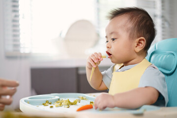 Asian little baby holding a baby corn. A son seat on a high chair in the kitchen at home. Finger...
