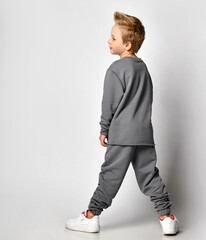 Little blond boy child with european appearance wearing trendy sportswear standing backside and looking aside on studio copy space demonstrating new clothing collection. Full-length portrait shot